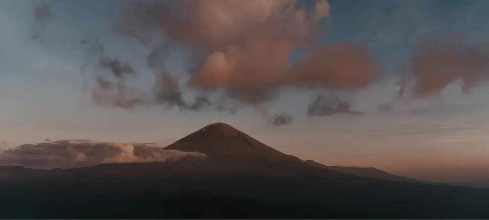 A distant view of Mount Meru rising above darkened foothills at dusk, with soft pink and gray clouds drifting across the sky and low clouds surrounding the mountain’s base.